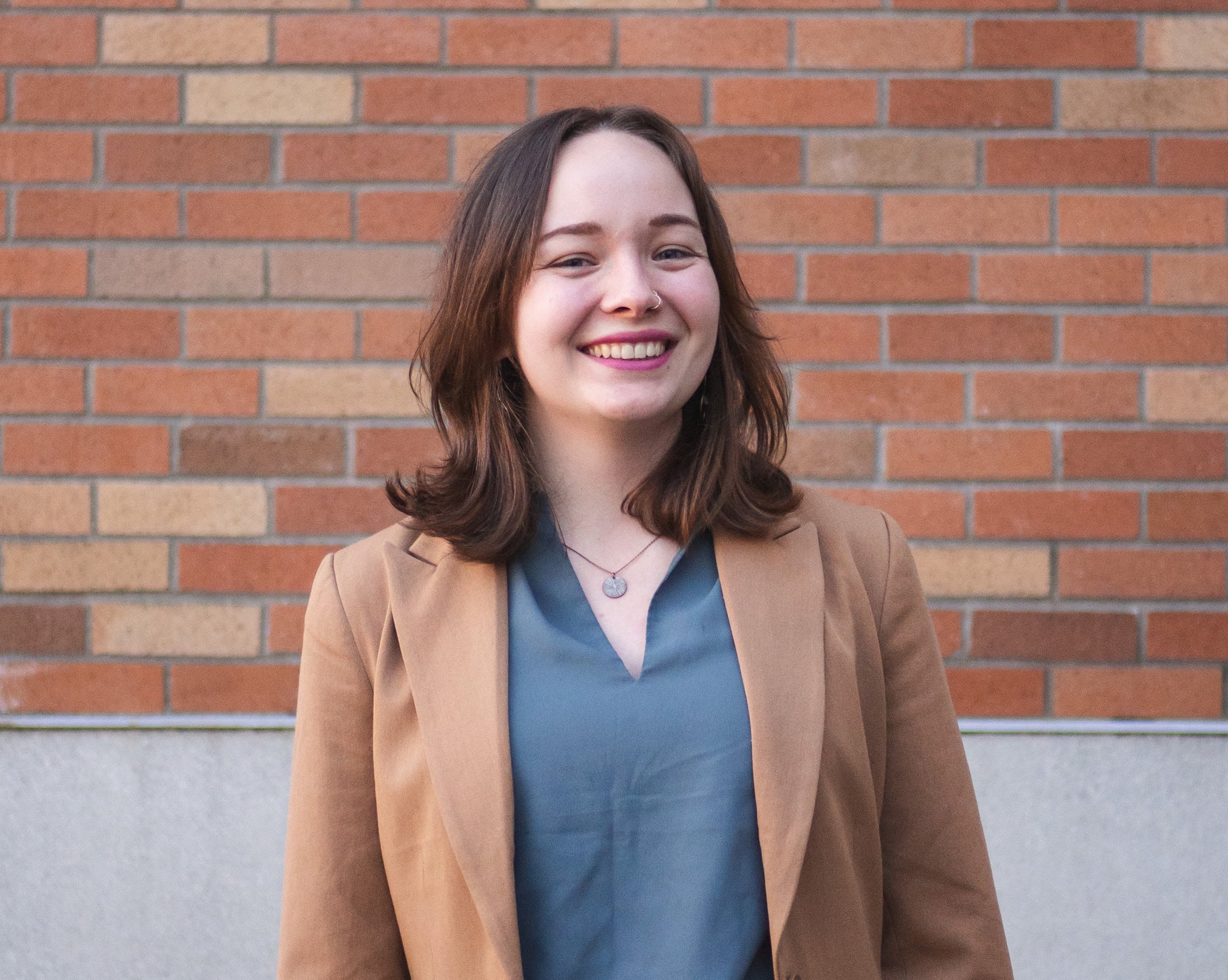 Photo of me in a blue-green shirt and brown blazer standing in front of a brick wall